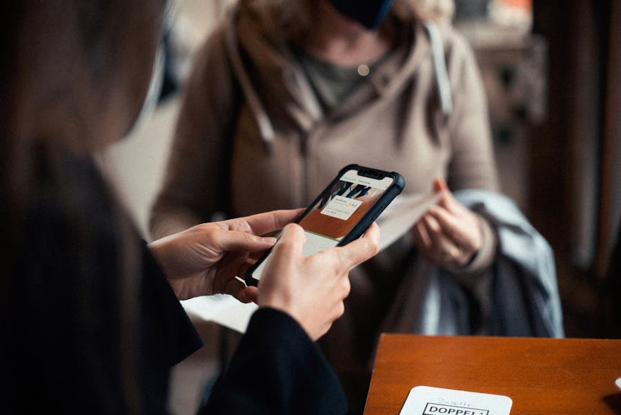 Uma pessoa escaneia um código QR usando um smartphone em um ambiente de evento. Ao fundo, um convidado segura um documento ou crachá, pronto para ser salvo. A cena ilustra um momento de check-in digitalizado, fluido e profissional, típico de um evento gerenciado com um aplicativo de controle de acesso.