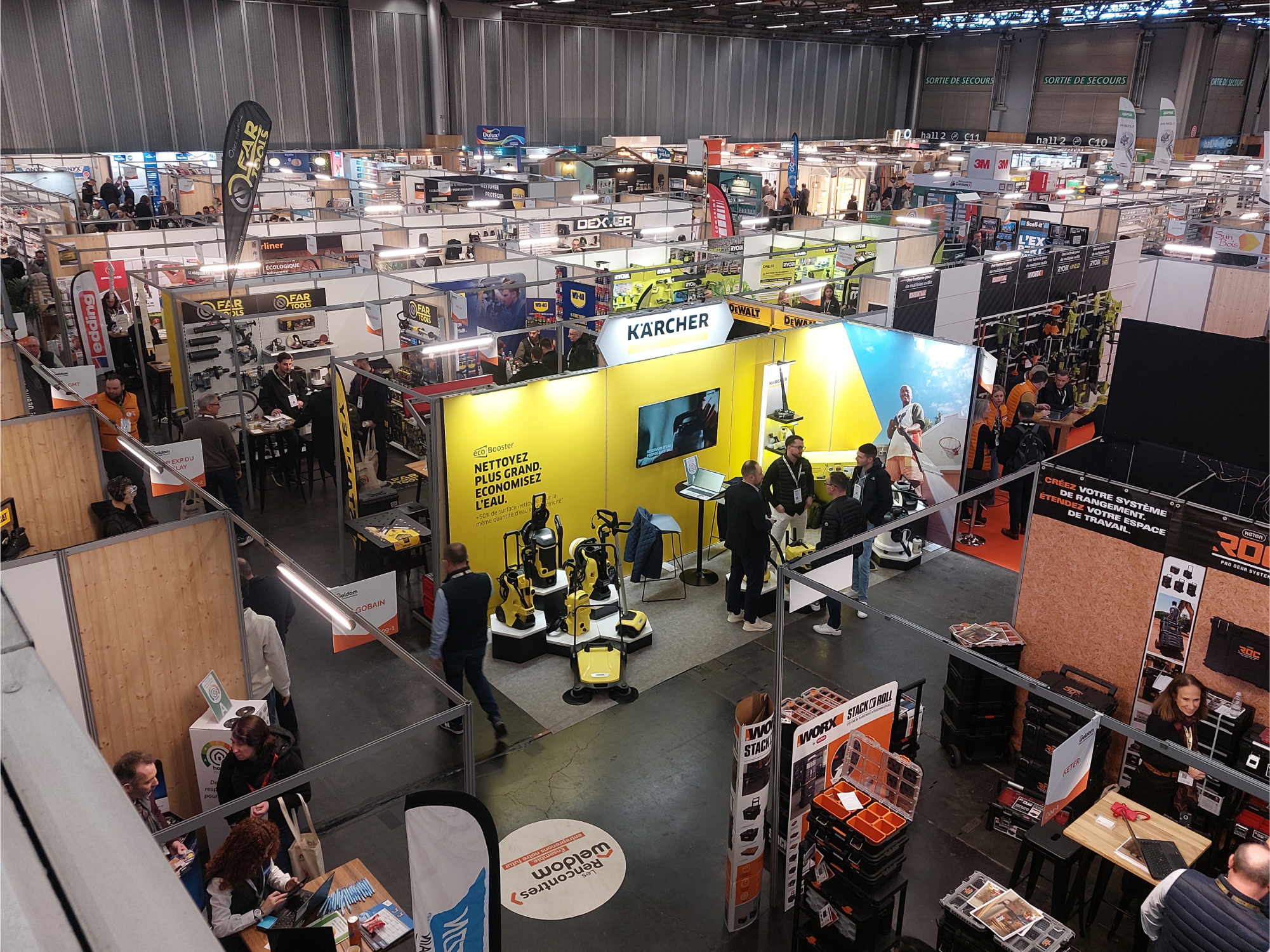 Vue aérienne d'un salon professionnel avec de nombreux stands d'exposants, dont Kärcher et d'autres marques du secteur industriel et de l'outillage. Les visiteurs échangent avec les exposants, explorent les produits exposés et circulent dans les allées du salon.