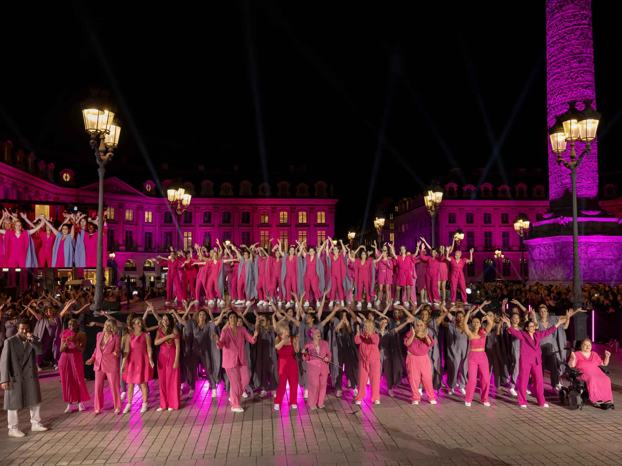 Une scène captivante lors d'Octobre Rose 2024, où une place publique est illuminée en rose pour soutenir la lutte contre le cancer du sein. Des participants vêtus de rose et de gris réalisent une danse synchronisée sous les lumières, symbolisant l'unité et l'engagement pour cette cause essentielle.