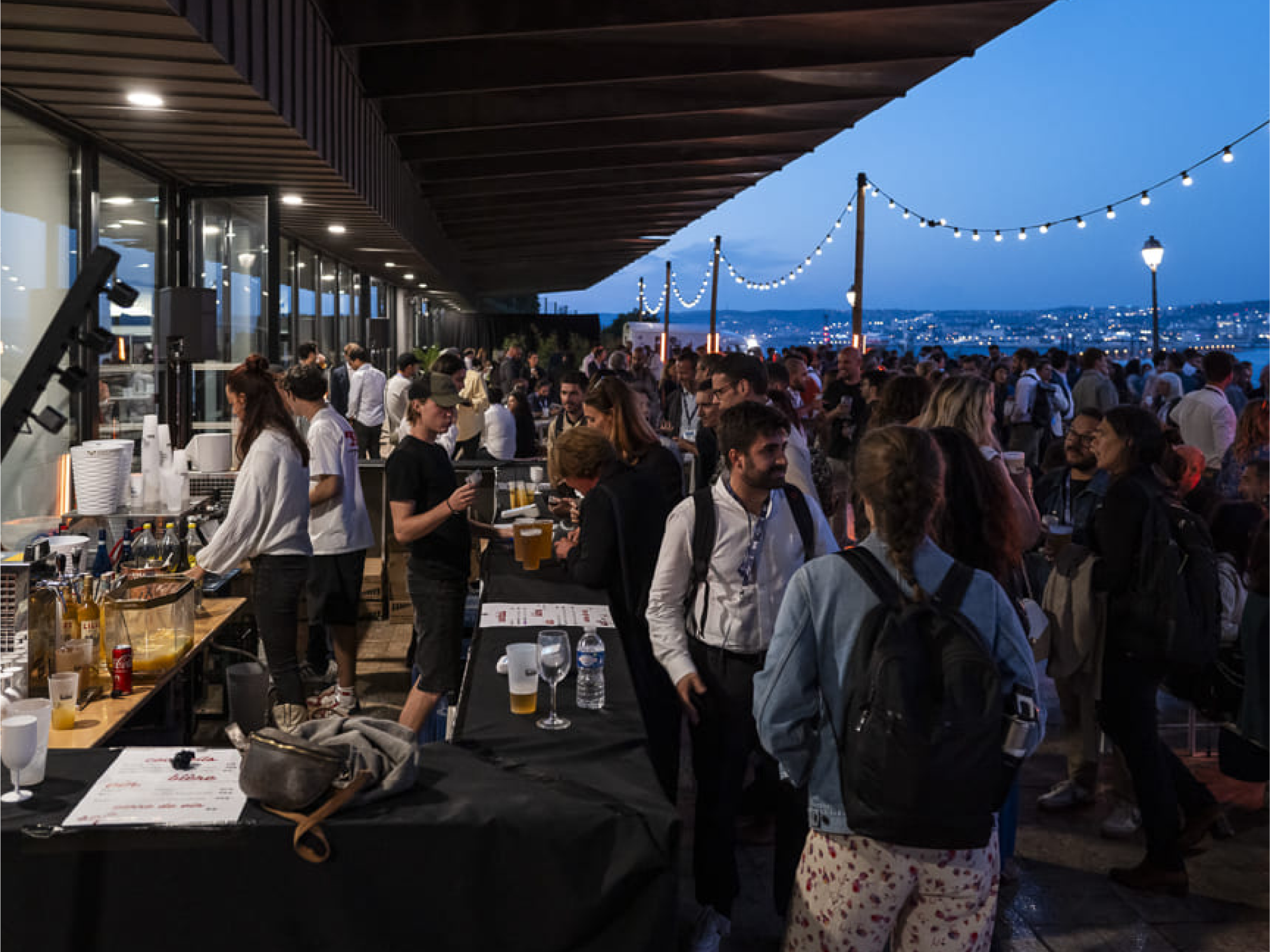 Soirée networking de la French Tech Aix-Marseille avec de nombreux participants réunis en extérieur sur une terrasse animée, ambiance conviviale avec bar, guirlandes lumineuses et vue sur la ville au crépuscule.