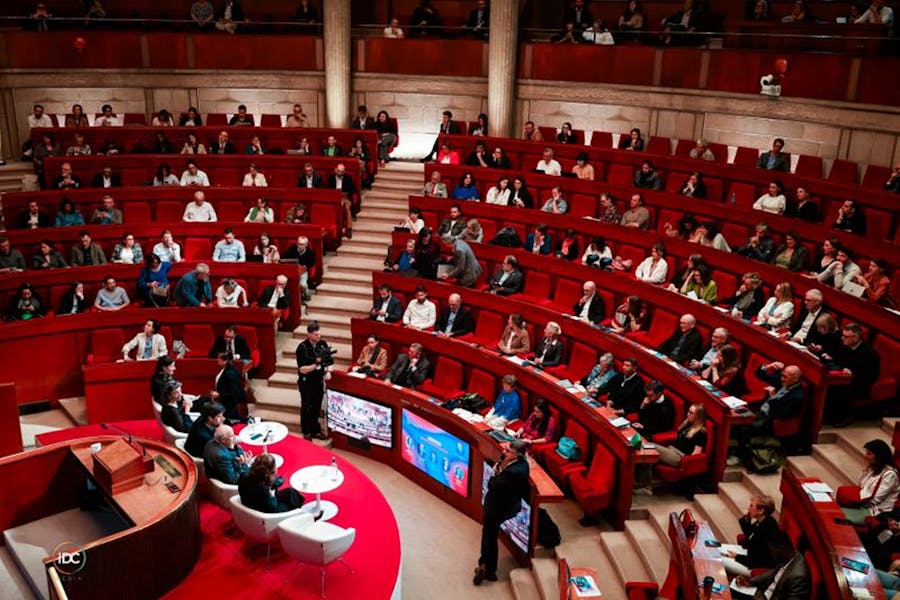 Vue d’ensemble d’un amphithéâtre circulaire rempli de participants assistant à une conférence. Au centre, plusieurs intervenants sont assis sur des fauteuils blancs, entourés d’écrans et de caméras. Le public est installé sur des gradins rouges.
