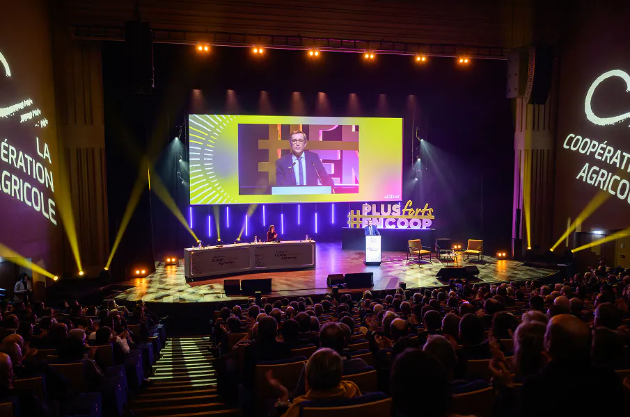 Vue d’un auditorium rempli de spectateurs assistant à une conférence, avec un intervenant à la tribune sur scène, un grand écran projetant son image, et des éclairages de scène colorés.