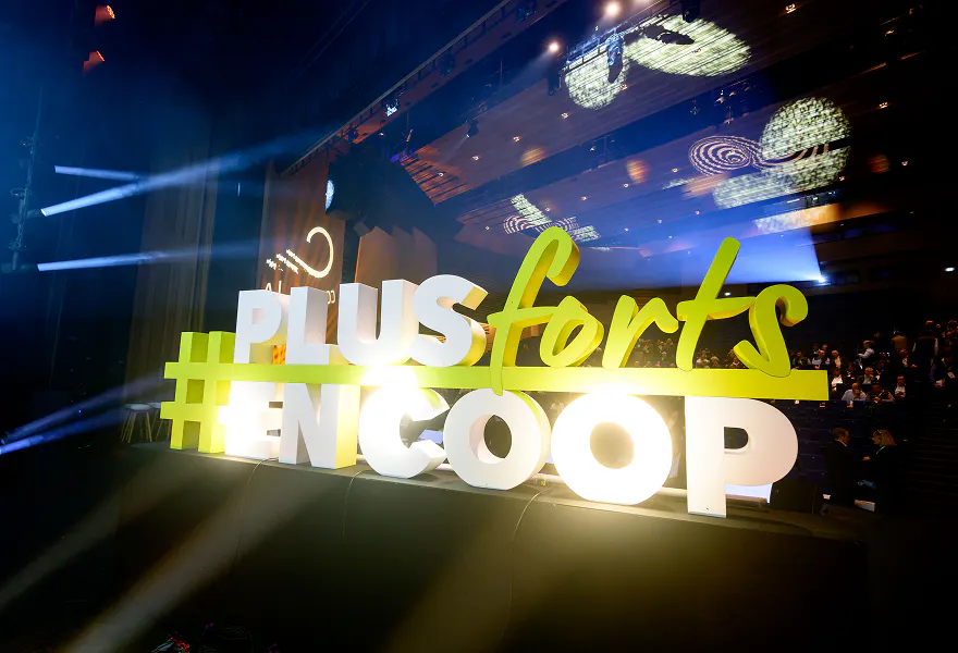Lettrage lumineux “#PLUSforts EN COOP” installé sur une scène avec éclairages colorés, dans une salle de conférence où des participants sont assis en arrière-plan.