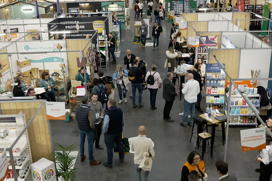 Vue en hauteur d’un salon professionnel intérieur avec plusieurs stands en bois alignés, où de nombreux visiteurs circulent, discutent et découvrent des produits exposés sur des présentoirs.