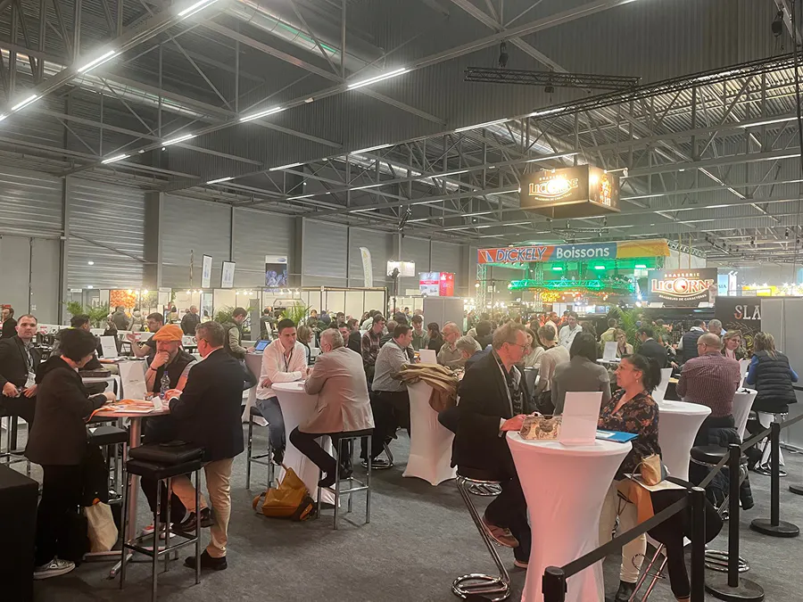 Espaço de evento com alta densidade de participantes participando de reuniões de negócios e networking, com múltiplas mesas e encontros simultâneos organizados em formato de business meetings.