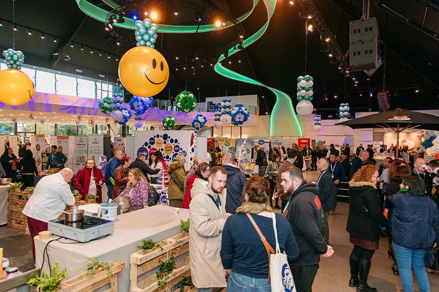 Vue d'ensemble de la salle d'exposition décorée de ballons et guirlandes, avec visiteurs, stands fournisseurs et espace démonstration culinaire.