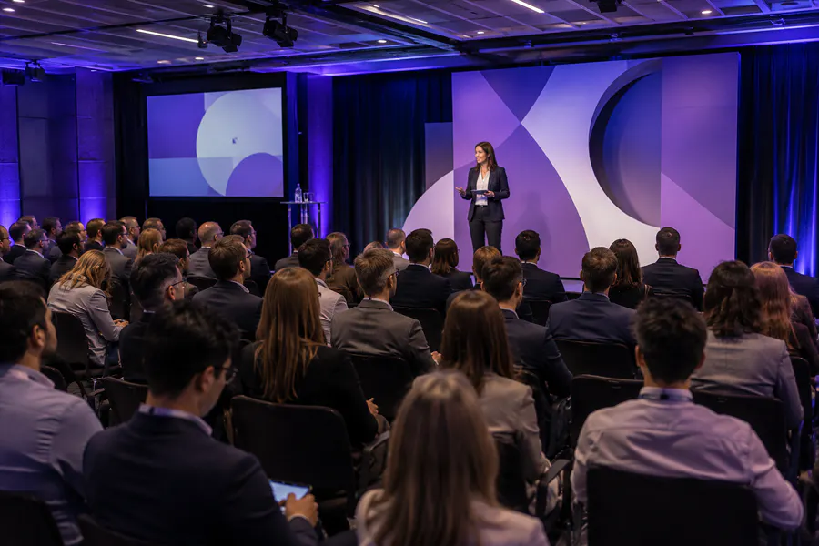 Photo of a conference in a plenary hall: a speaker addresses the audience on stage in front of a designer backdrop in purple tones, with a large seated audience listening attentively in a dim blue-purple lighting atmosphere.