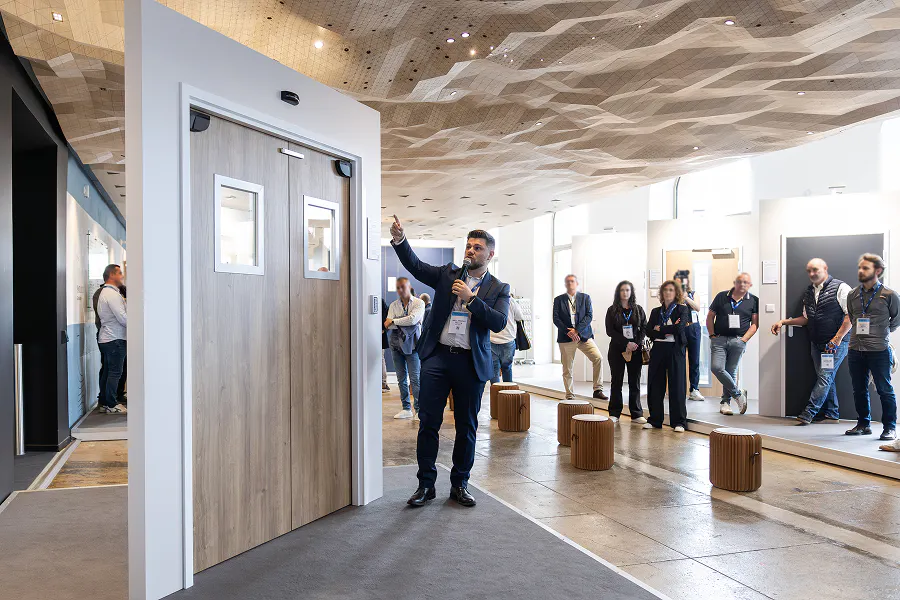 Homme en costume présentant une porte en bois à un groupe de visiteurs dans un espace d'exposition au plafond sculpté.