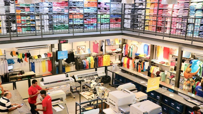 Interior da loja da Dimona na Barra da Tijuca com várias camisas coloridas e máquinas de estamparia