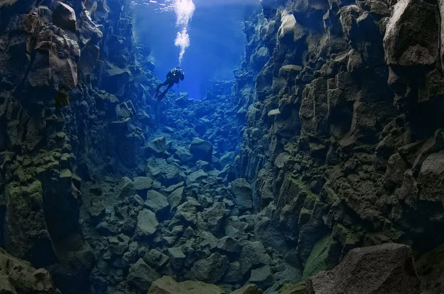 a crevice underwater with rocks walls and floor and a scuba diver in the distance