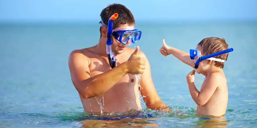 a man and child, each with a mask and snorkel on giving each other thumbs up in shallow open water