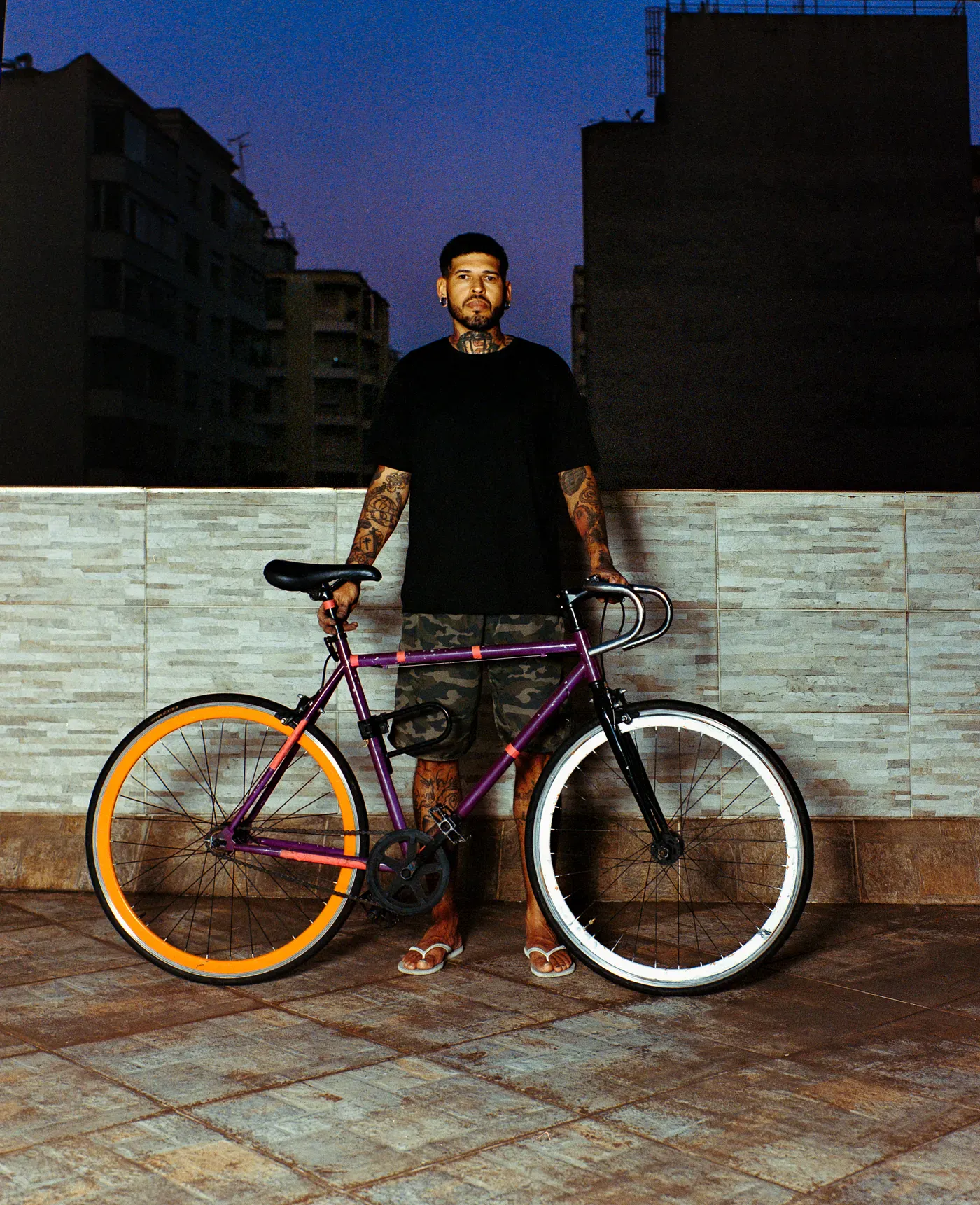 A tattooed man in a black shirt stands outdoors at dusk holding a purple bicycle with an orange rear wheel and a silver front wheel. Silhouetted city buildings are visible against the dark blue sky behind him.