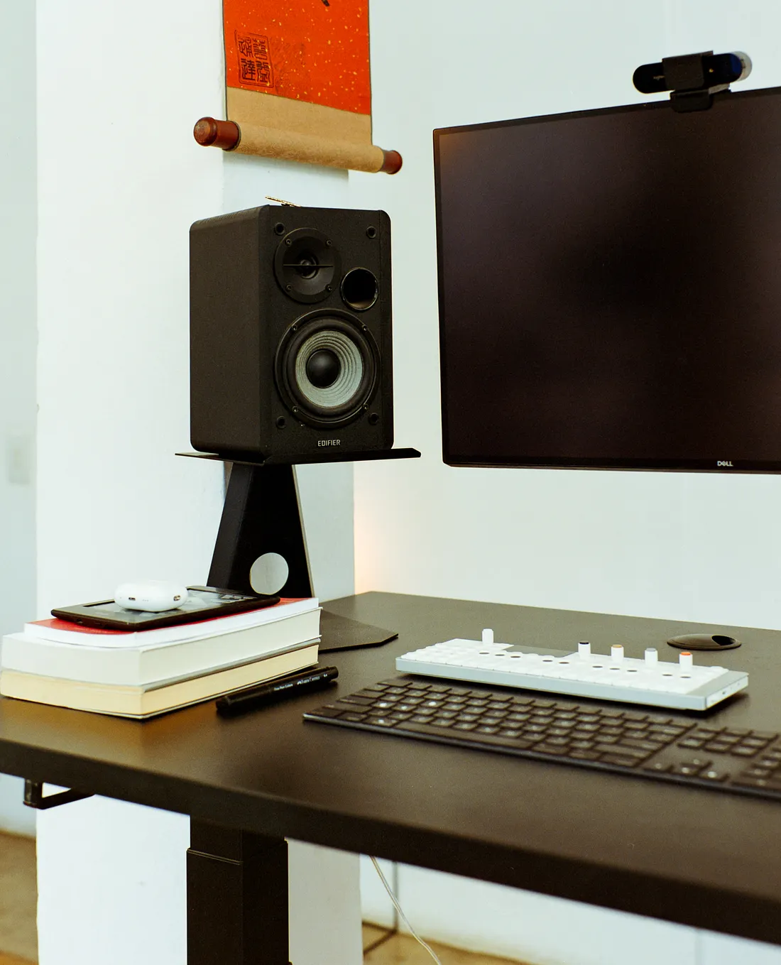 A minimalist desk setup with a dark tabletop featuring a computer keyboard, a compact white synthesizer, a stack of books, an Edifier speaker on a black stand, and a Dell monitor with a top-mounted webcam against a white wall.