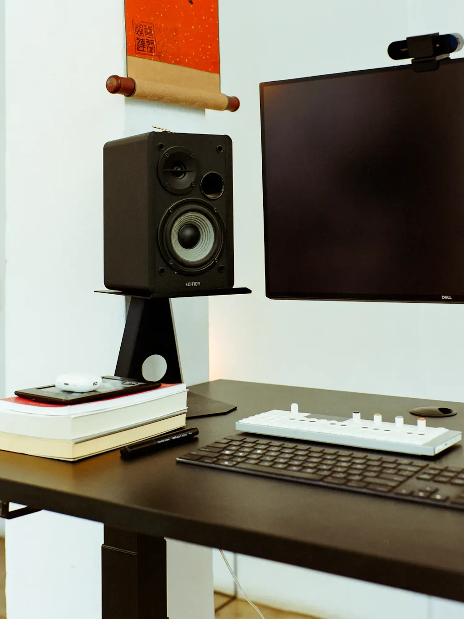 A minimalist desk setup with a dark tabletop featuring a computer keyboard, a compact white synthesizer, a stack of books, an Edifier speaker on a black stand, and a Dell monitor with a top-mounted webcam against a white wall.