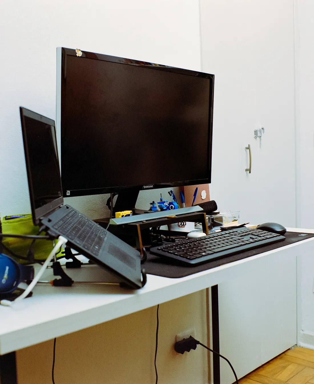 A white desk features a desktop monitor, a laptop on an angled stand, a wireless keyboard, and a mouse. Blue over-ear headphones, a pen cup, and a small calendar are arranged on the left side.