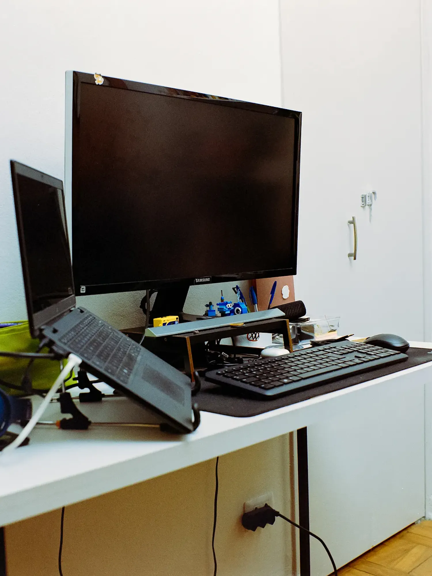 A white desk features a desktop monitor, a laptop on an angled stand, a wireless keyboard, and a mouse. Blue over-ear headphones, a pen cup, and a small calendar are arranged on the left side.