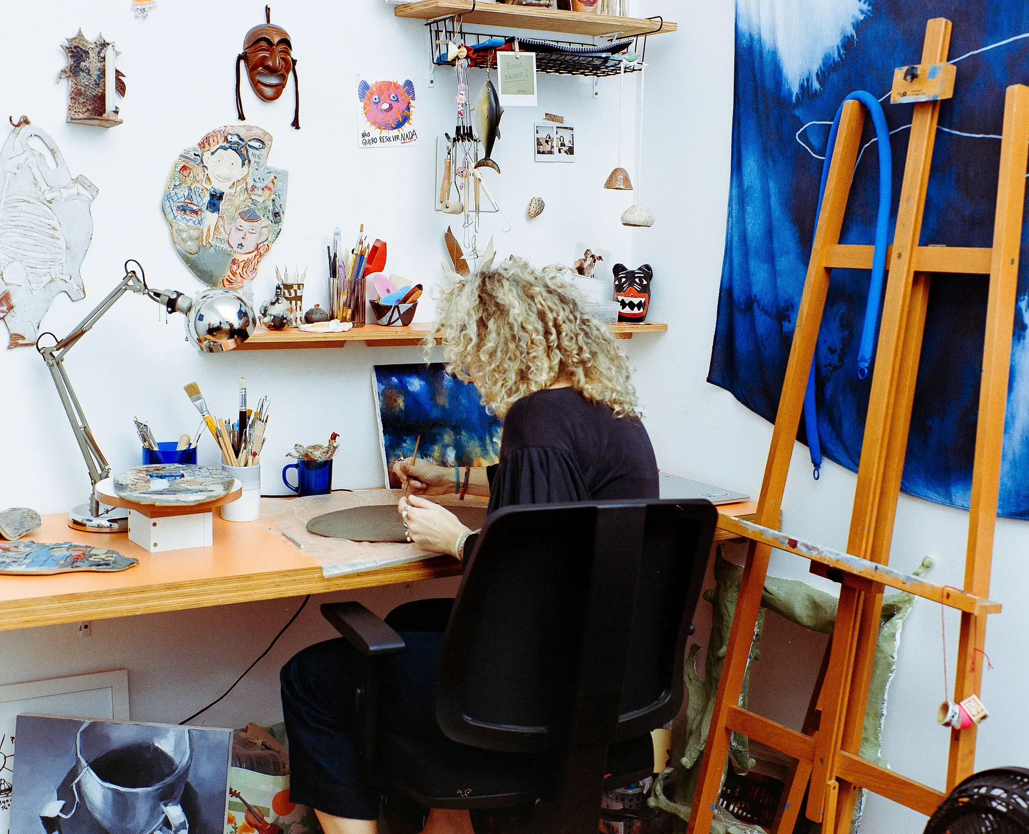 An artist with curly hair sits at an orange desk working on a piece of clay. The art studio is filled with creative tools, a wooden easel, wall decorations, shelves with supplies, and a large blue fabric hanging on the wall.