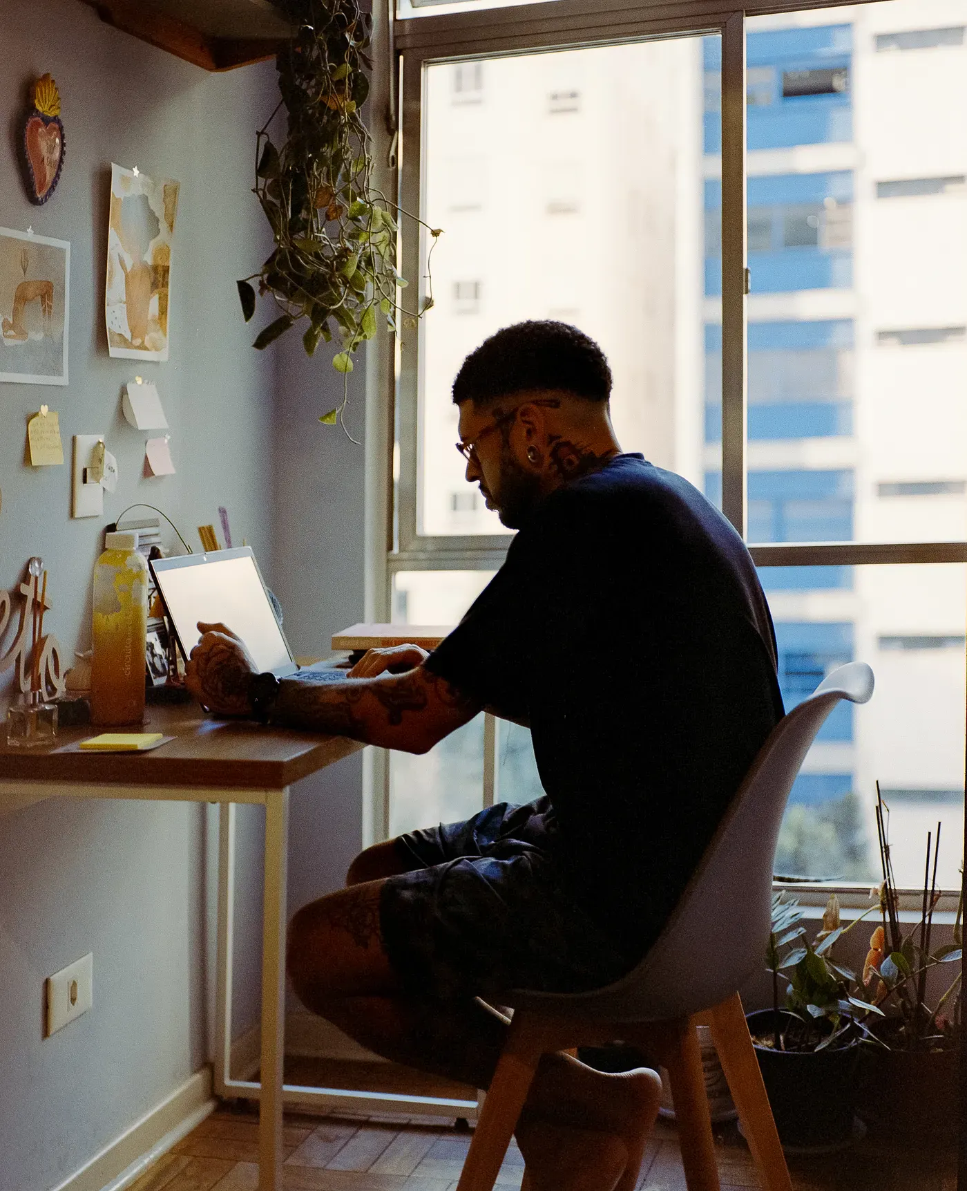 A tattooed man sits at a small wooden desk working on an open laptop. He faces a large window overlooking city buildings, with a hanging plant above and various notes on the wall to his left.