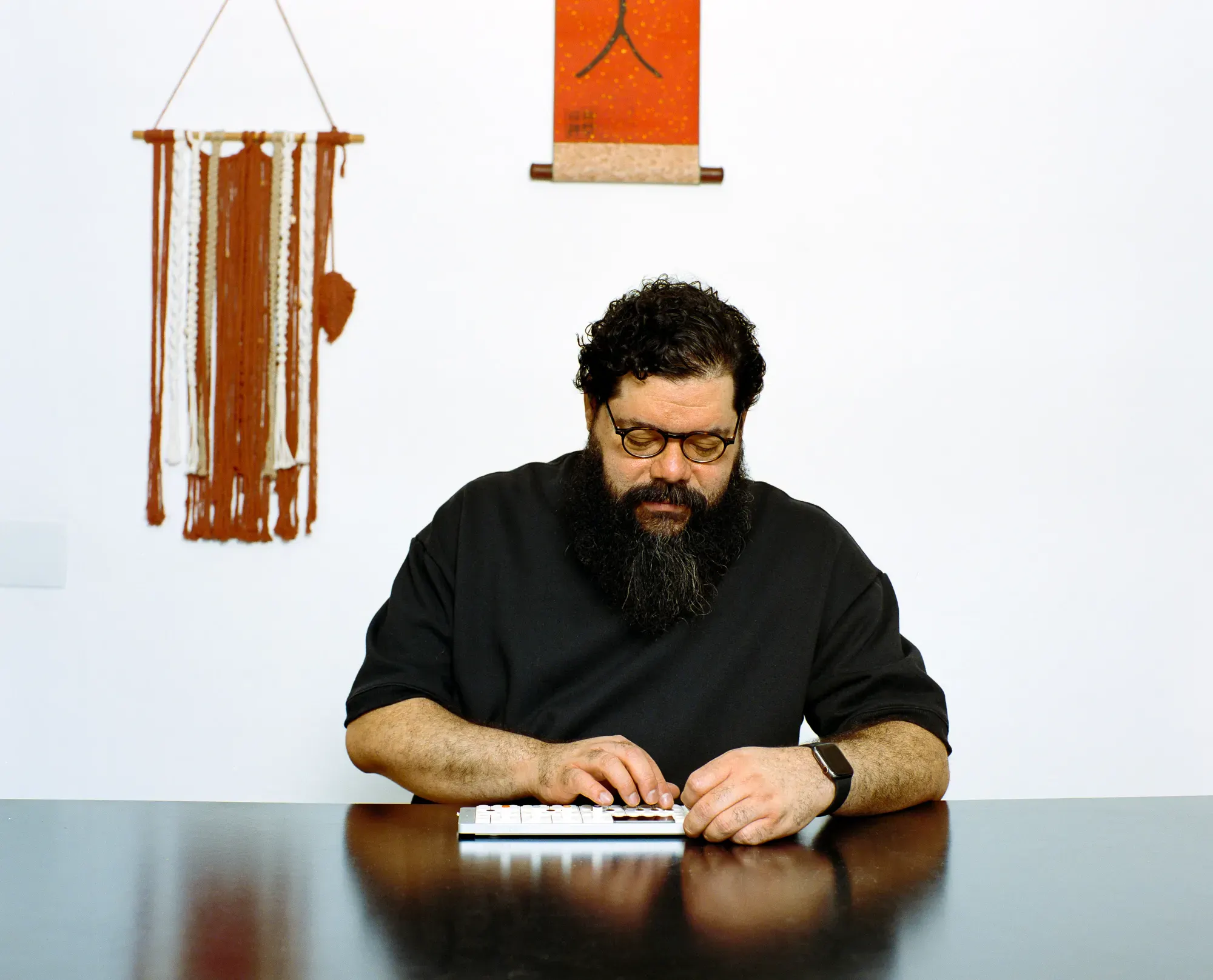 A man with a full beard, curly hair, and glasses sits at a dark table, looking down while playing a compact white synthesizer. A woven macrame wall hanging and a small orange scroll hang on the white wall behind him.