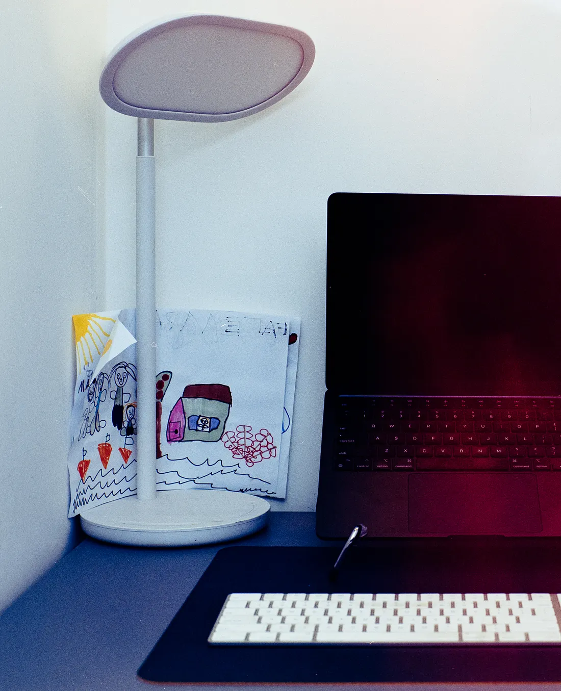 A dark laptop sits open on a stand on a desk, positioned between a white desk lamp with a child's drawing behind it, and a second computer monitor. A white wireless keyboard rests on a black desk mat in the foreground.