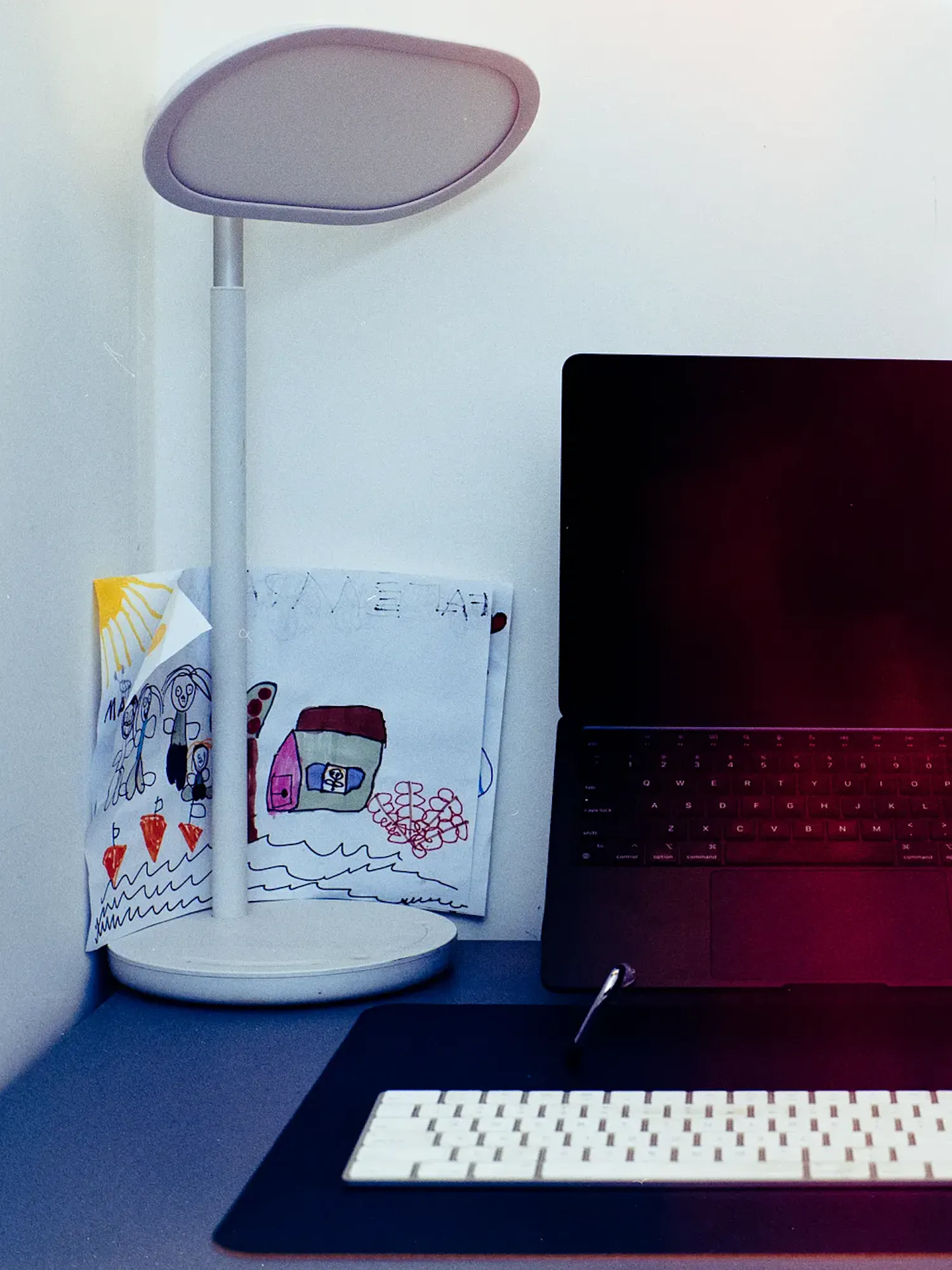A dark laptop sits open on a stand on a desk, positioned between a white desk lamp with a child's drawing behind it, and a second computer monitor. A white wireless keyboard rests on a black desk mat in the foreground.