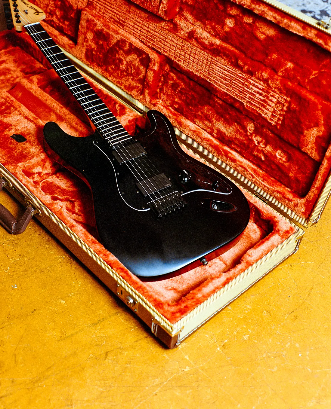 An all-black Fender Stratocaster electric guitar nestled inside an open hardshell case lined with bright orange plush fabric, resting on a textured golden-yellow floor.