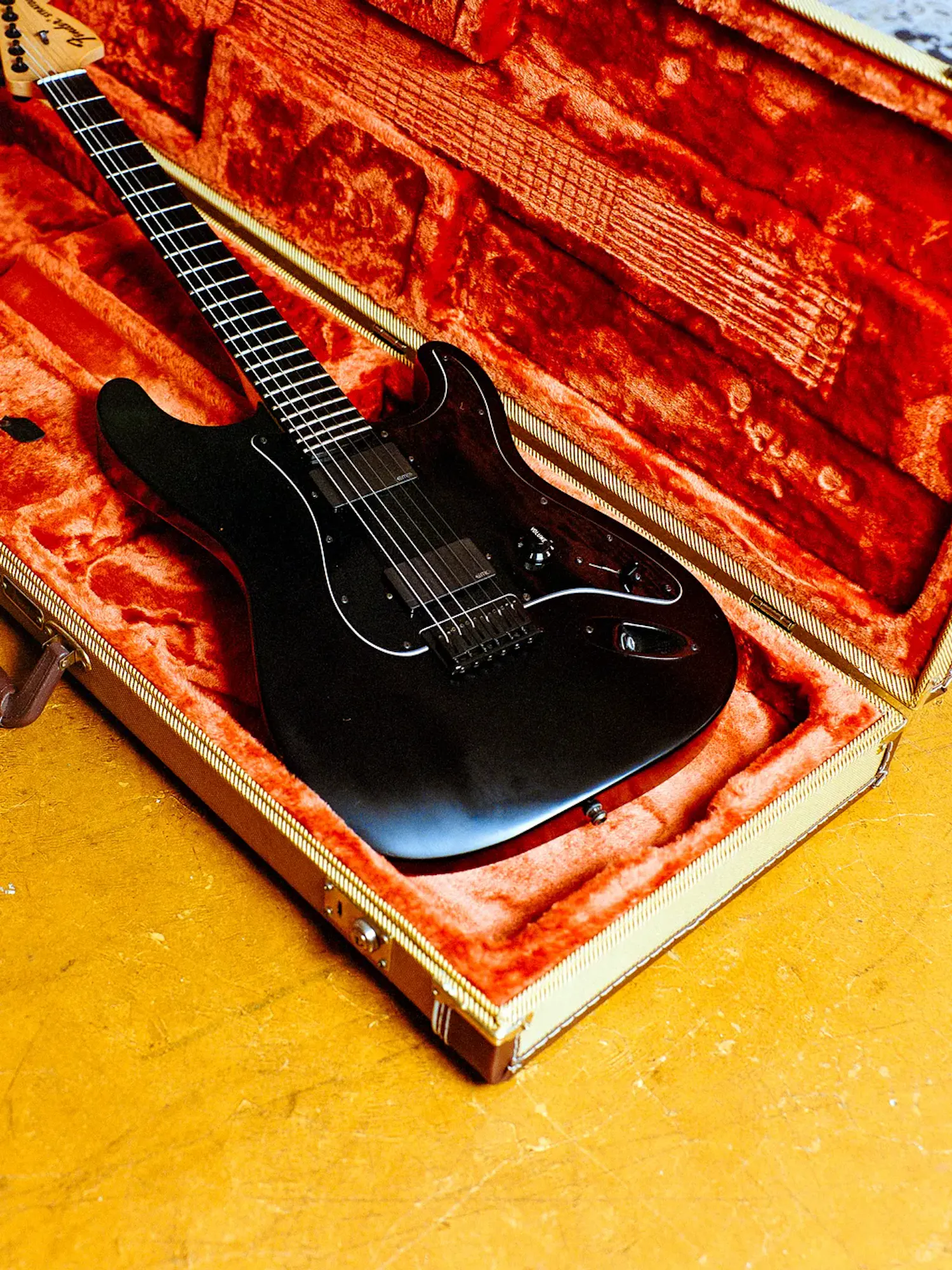 An all-black Fender Stratocaster electric guitar nestled inside an open hardshell case lined with bright orange plush fabric, resting on a textured golden-yellow floor.