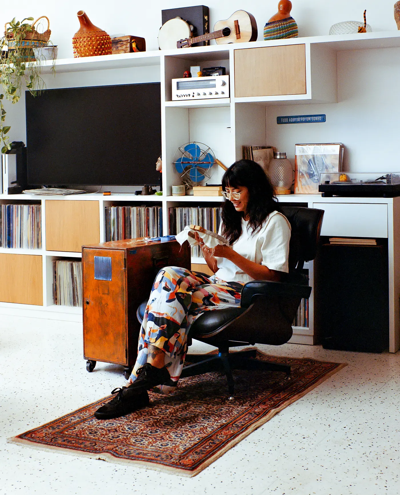 A smiling woman sits in a black lounge chair on a patterned rug, working on embroidery. Behind her is a white wall unit filled with vinyl records, a television, a record player, instruments, and various decorative items.