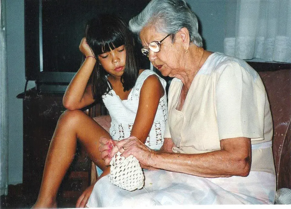 An elderly woman with glasses and short grey hair sits crocheting a white yarn piece. Next to her, a young girl with dark hair and bangs rests her head on her hand, looking down intently at the woman's hands working.