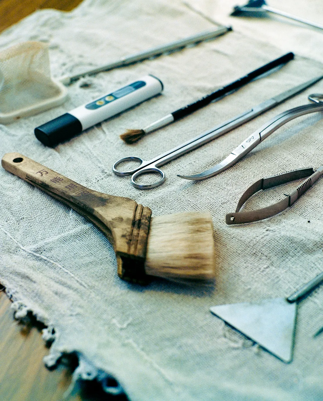 Various aquascaping tools, including stainless steel curved scissors, tweezers, a triangle sand flattener, a wooden-handled paintbrush, and a digital water tester, are neatly arranged on a textured light cloth.