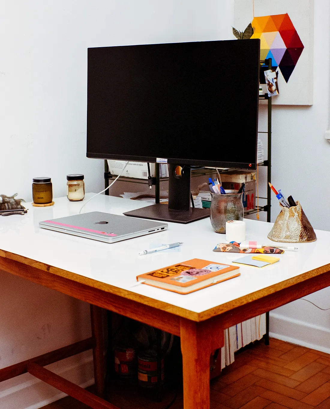 A wooden desk with a white top holds a large black computer monitor, a closed silver laptop, an orange notebook, and pen holders. In the background, a colorful geometric artwork hangs on a white wall.