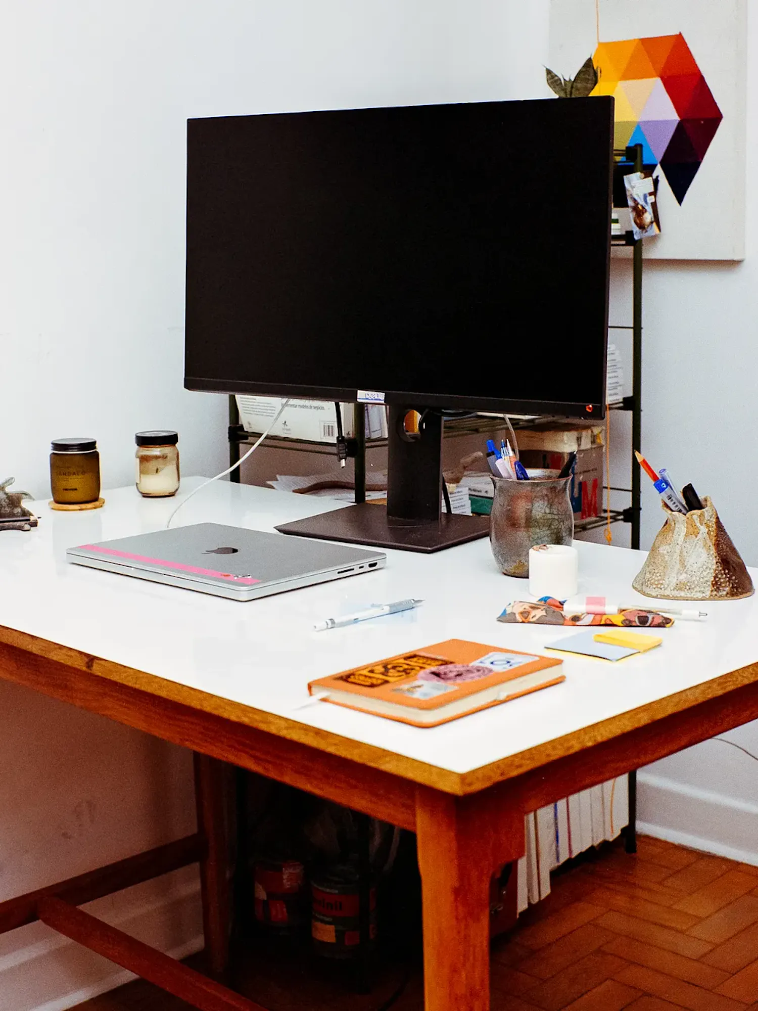 A wooden desk with a white top holds a large black computer monitor, a closed silver laptop, an orange notebook, and pen holders. In the background, a colorful geometric artwork hangs on a white wall.