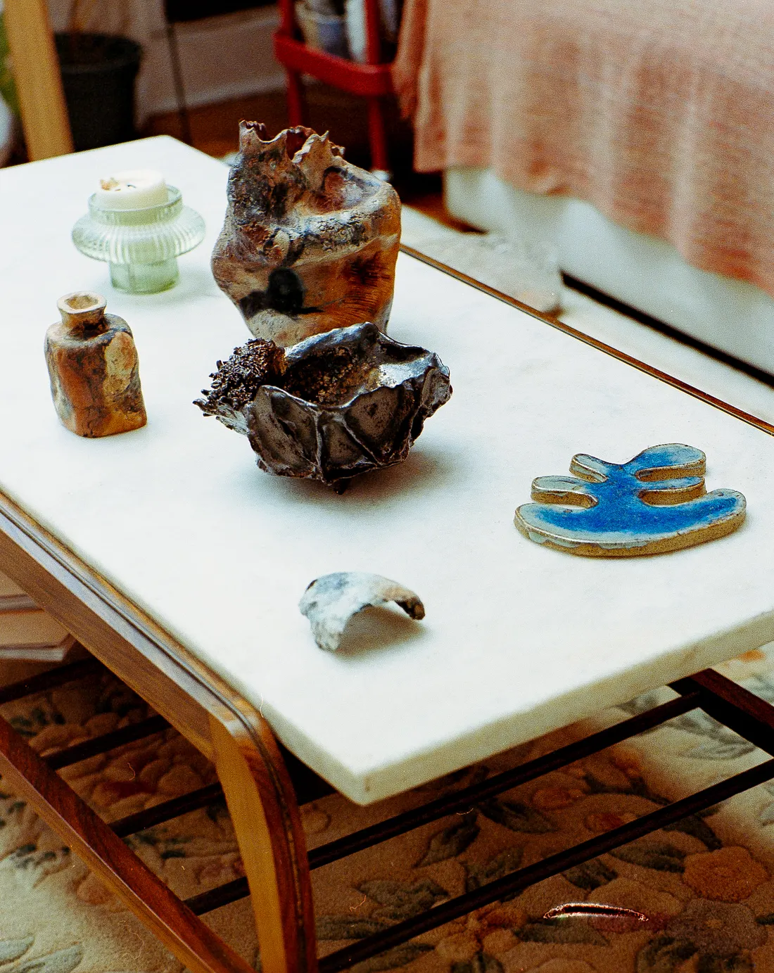 Various textured ceramic sculptures, a small vase, a blue glazed piece, and a candle sit on a white marble-top coffee table with a wooden frame.