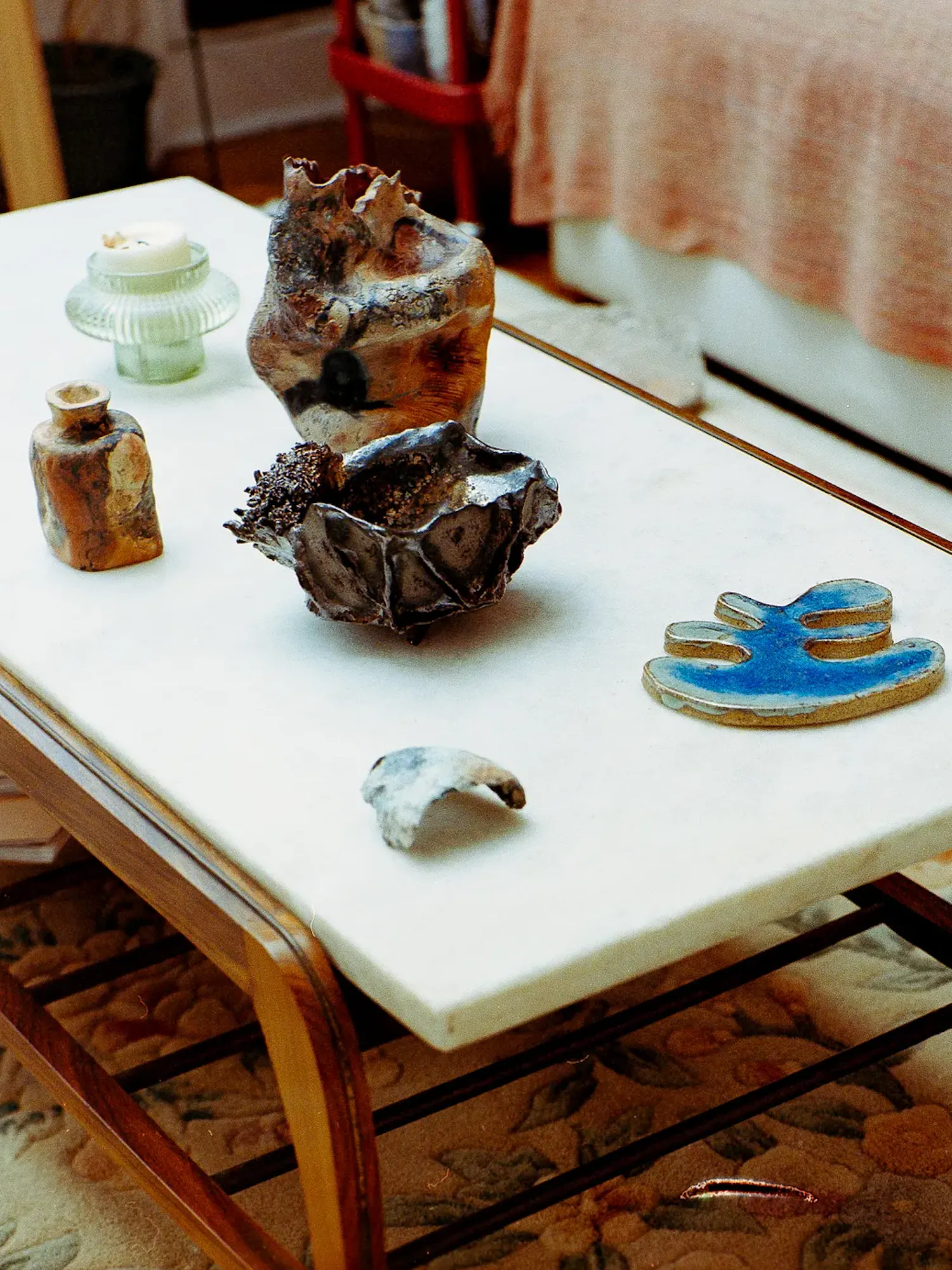 Various textured ceramic sculptures, a small vase, a blue glazed piece, and a candle sit on a white marble-top coffee table with a wooden frame.