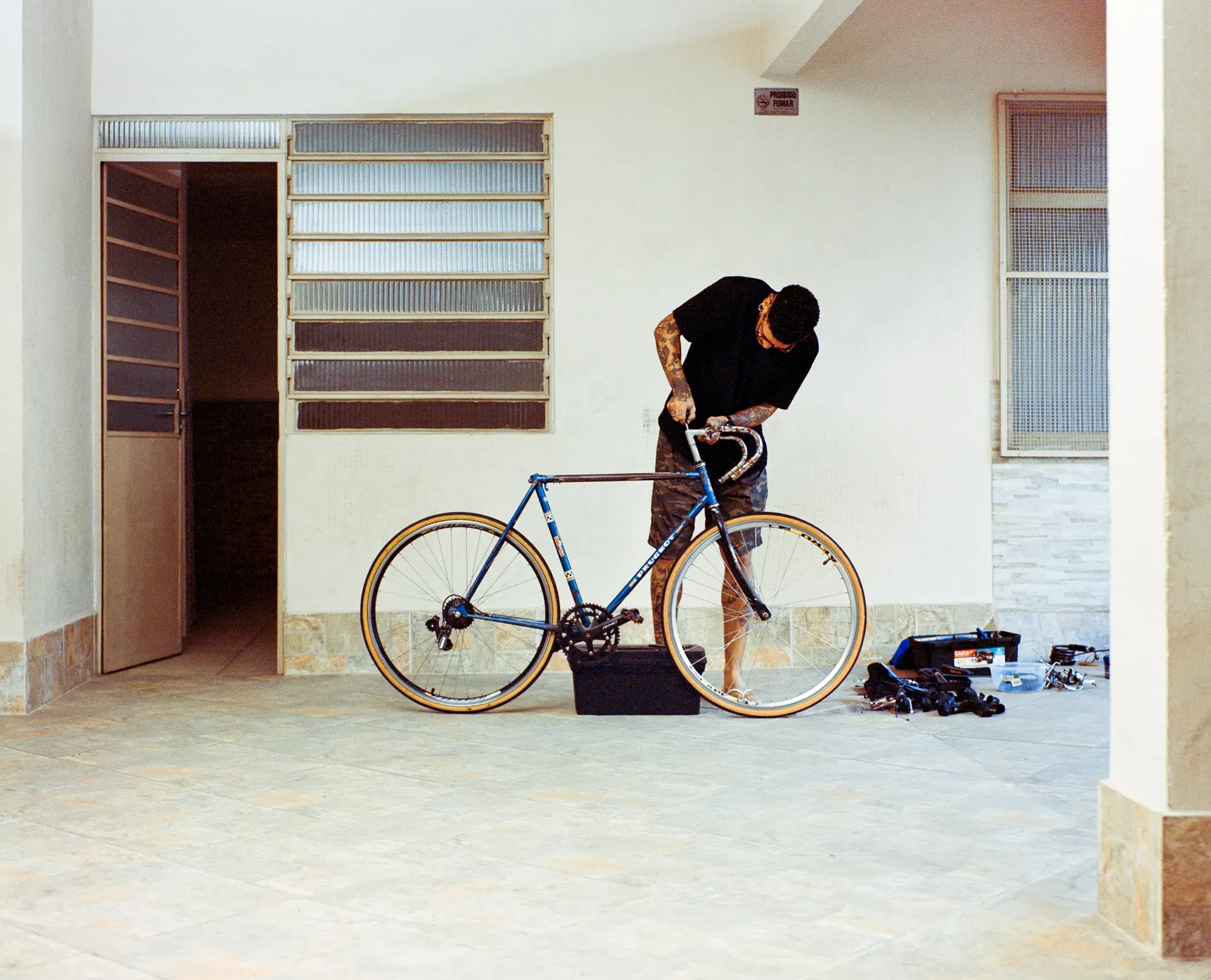 A tattooed man in a black shirt and camo shorts stands on an outdoor patio working on a blue Peugeot bicycle. Disassembled parts are clustered on the ground to his right.