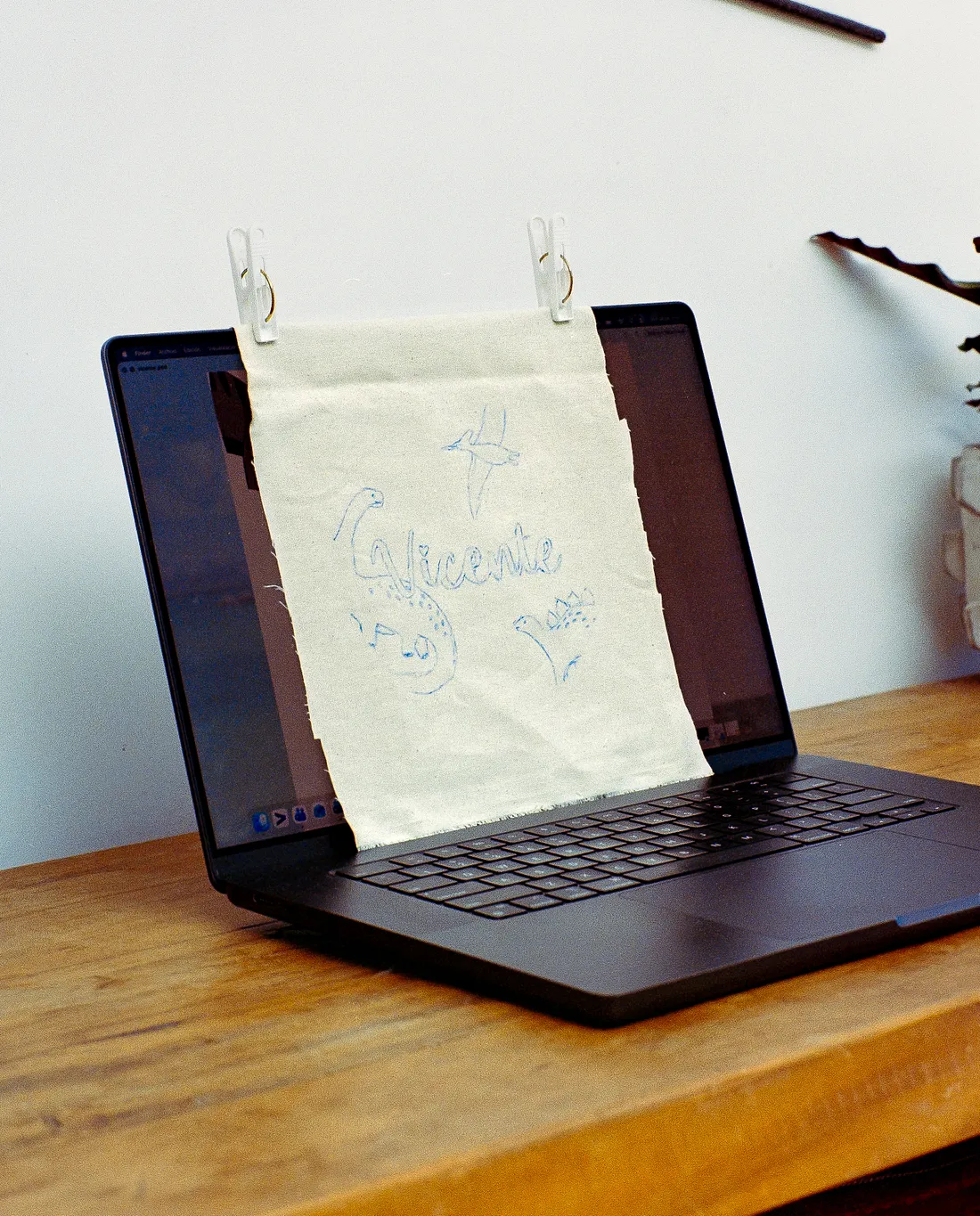 A light fabric sheet is clipped to an open laptop screen with two white clips. The fabric has blue hand-drawn sketches of dinosaurs and the name "Vicente." The laptop sits on a wooden table.