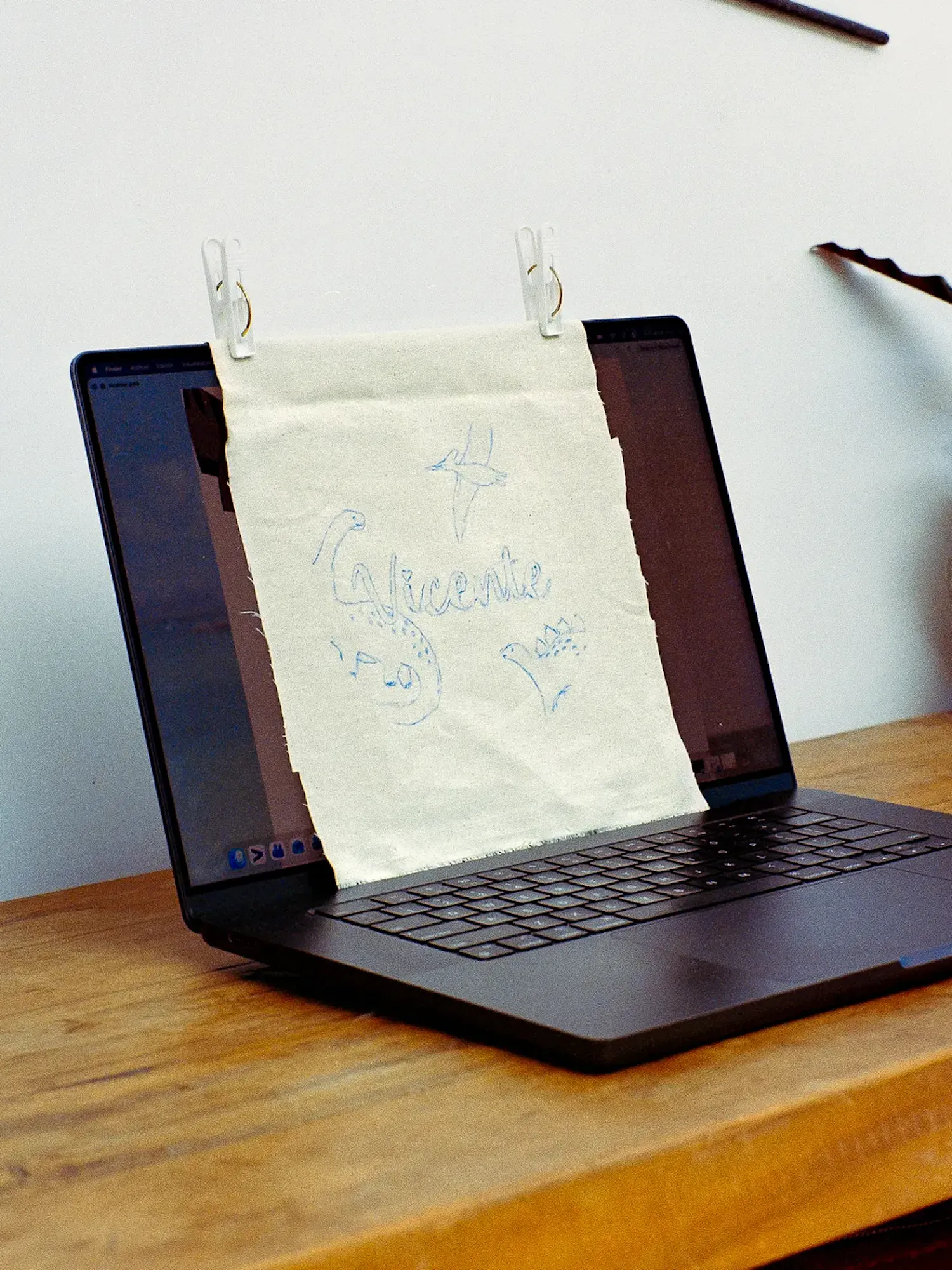 A light fabric sheet is clipped to an open laptop screen with two white clips. The fabric has blue hand-drawn sketches of dinosaurs and the name "Vicente." The laptop sits on a wooden table.