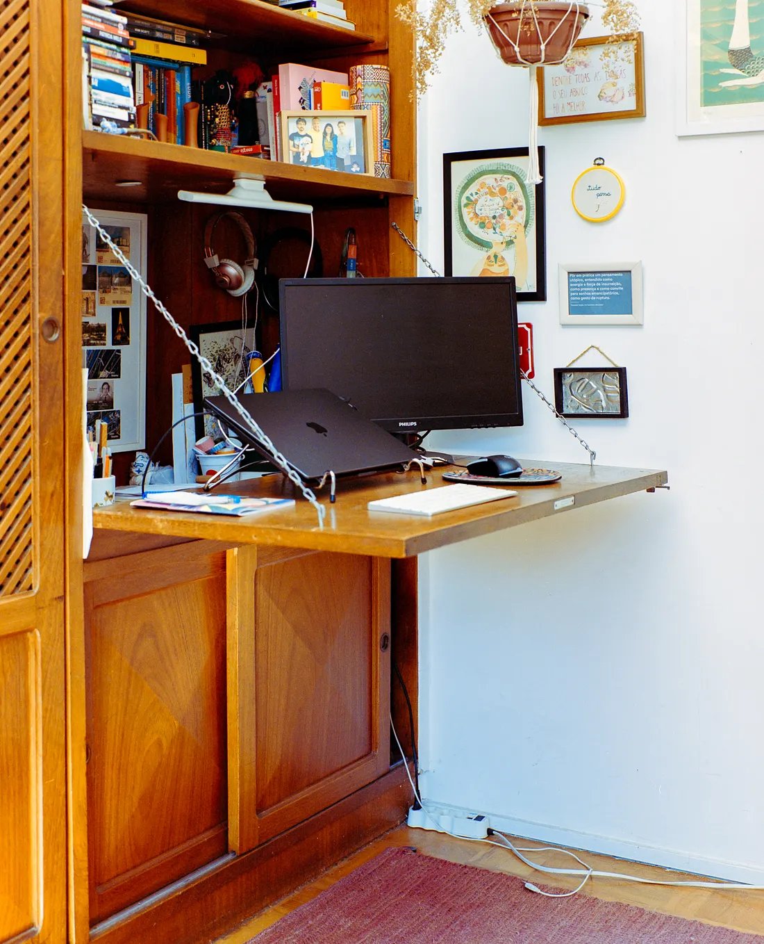 A wooden drop-down secretary desk holds a monitor, a laptop on a stand, a keyboard, and a mouse. The surrounding space features a built-in bookshelf filled with items, alongside several framed pictures on a white wall.