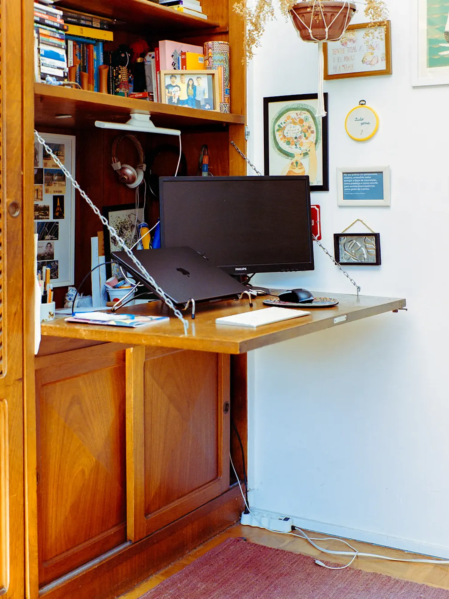 A wooden drop-down secretary desk holds a monitor, a laptop on a stand, a keyboard, and a mouse. The surrounding space features a built-in bookshelf filled with items, alongside several framed pictures on a white wall.