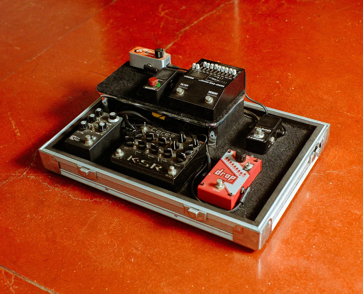 An organized guitar pedalboard in a silver hardshell case on an orange floor. It contains several effects pedals, including a black KSR Ceres preamp, a red DigiTech Drop pedal, a Boss EQ-200, and a TC Electronic PolyTune.