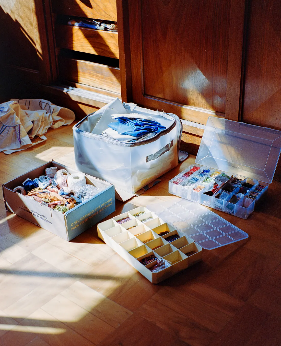 An assortment of craft supplies sits on a wooden floor in the sunlight. Items include a cardboard box filled with colorful embroidery floss and yarn, a fabric bin with blue cloth, and two plastic organizers filled with beads and threads.