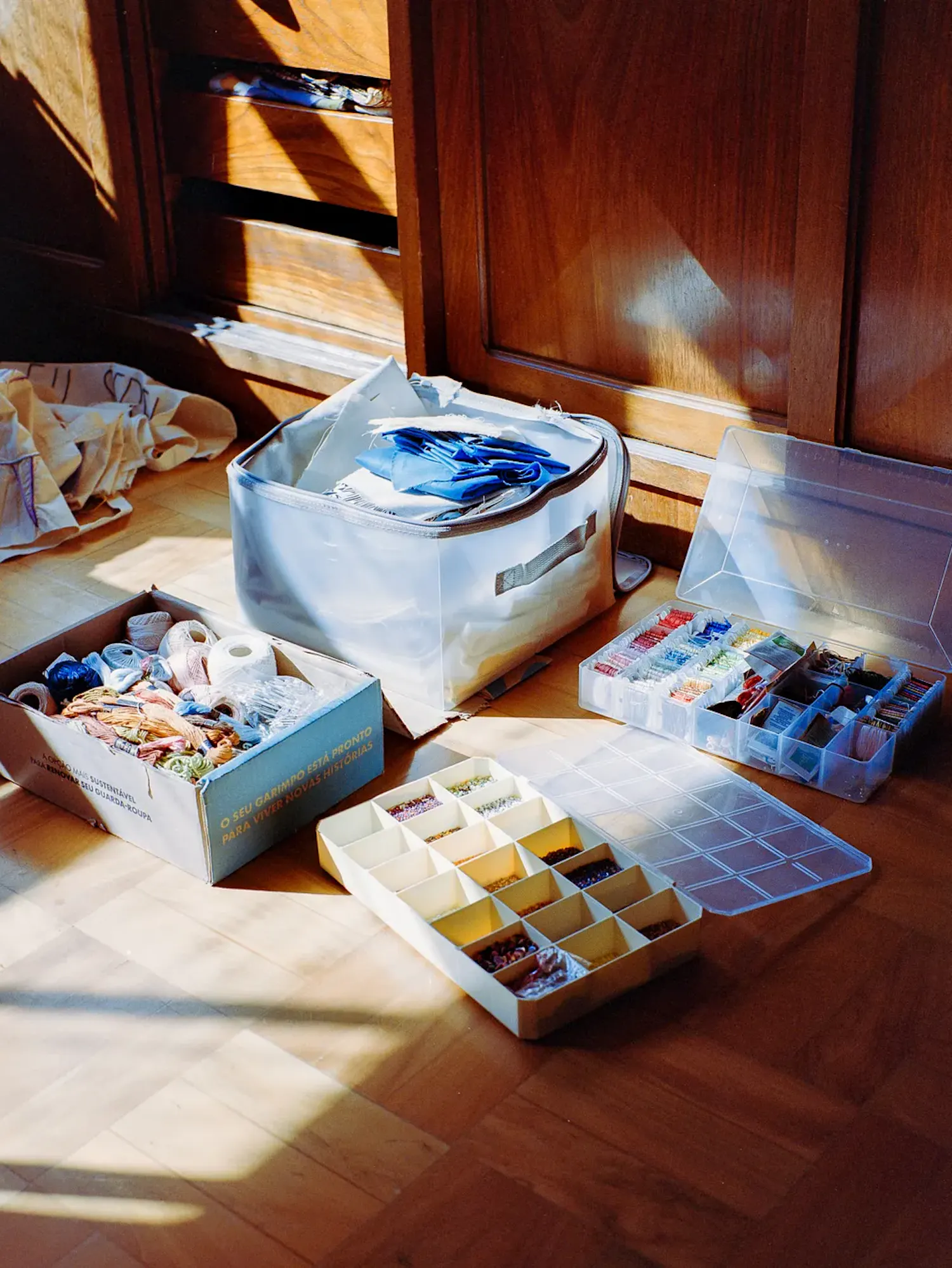 An assortment of craft supplies sits on a wooden floor in the sunlight. Items include a cardboard box filled with colorful embroidery floss and yarn, a fabric bin with blue cloth, and two plastic organizers filled with beads and threads.