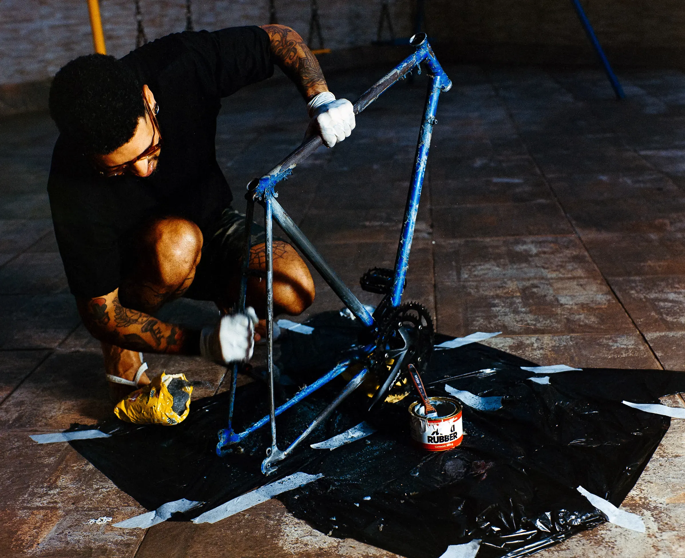 A tattooed man wearing gloves crouches on an outdoor tiled patio, working on a bare, blue bicycle frame placed over a black plastic sheet. A small can labeled "RUBBER" sits nearby with a brush inside.