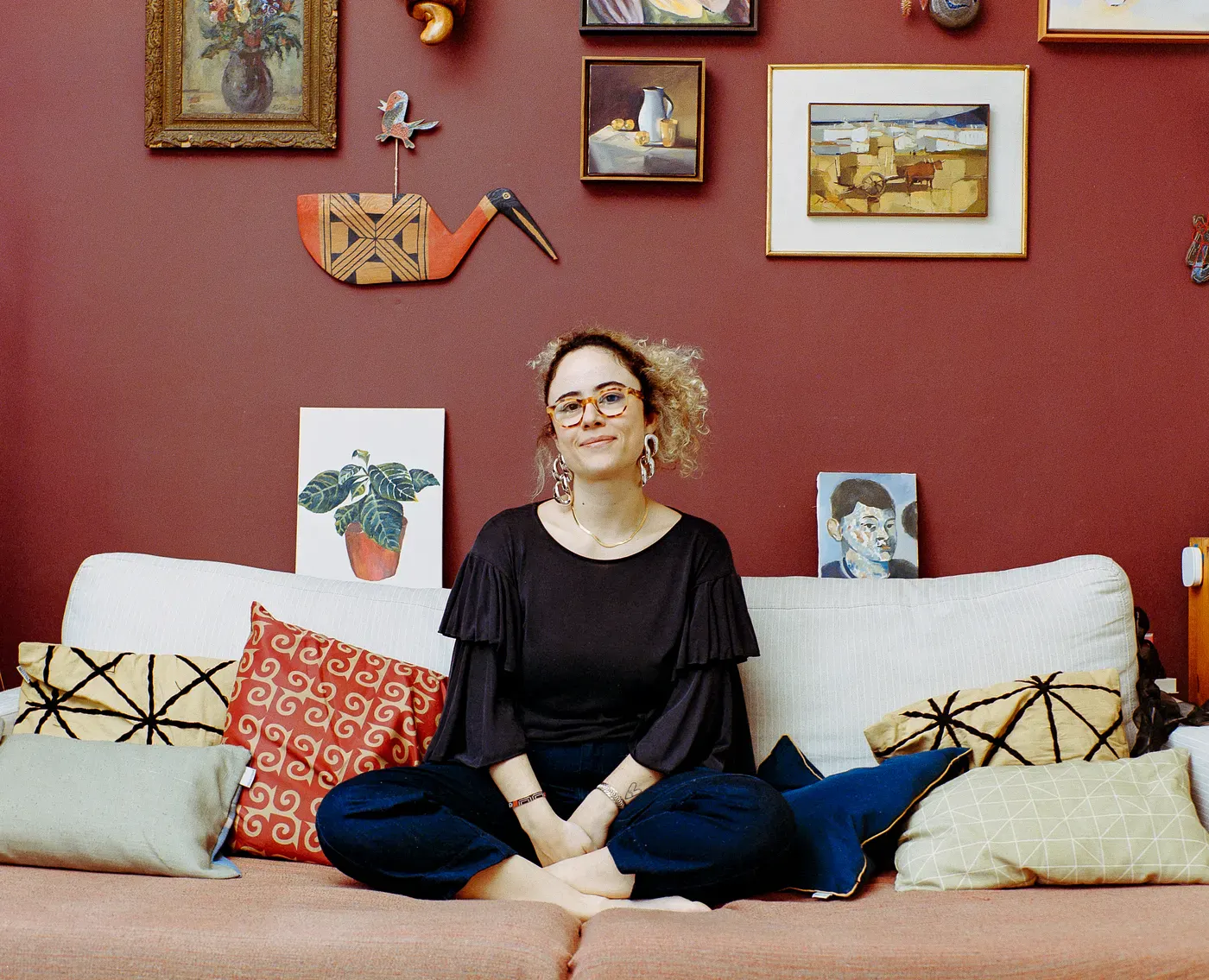 A woman with curly hair and glasses sits cross-legged on a couch with patterned pillows. Behind her, a dark red wall is decorated with various framed paintings and art pieces, including a bird sculpture and a plant painting.