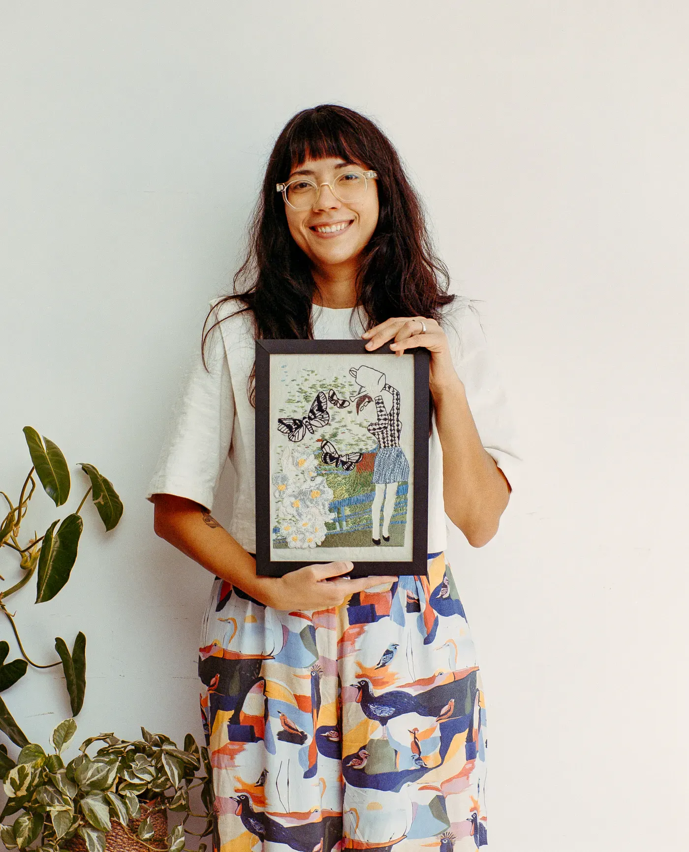A smiling woman with glasses and dark hair holds a framed embroidery piece depicting a person, butterflies, and flowers. She wears a white top and colorful bird-patterned pants against a plain white wall.