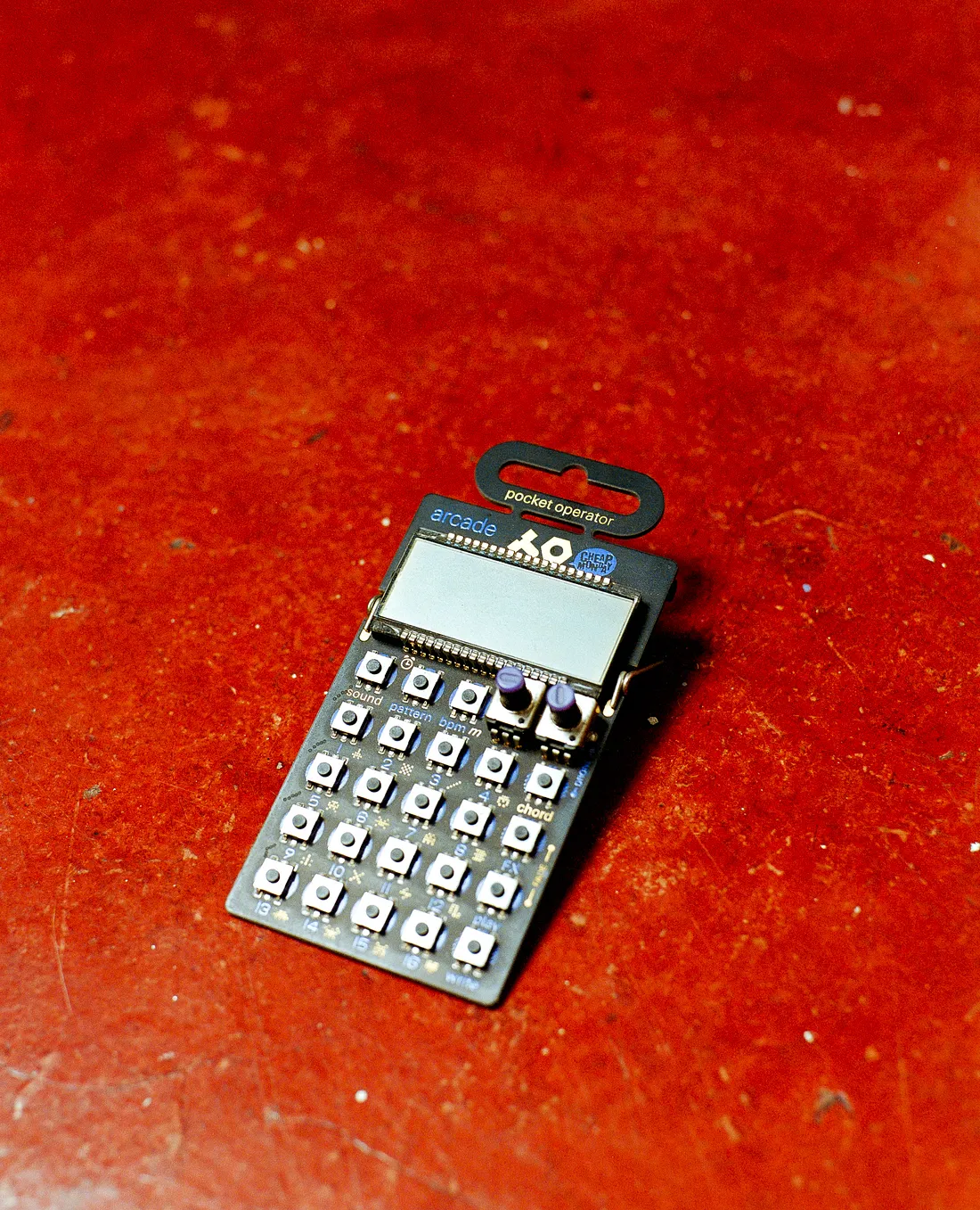 A small Teenage Engineering Pocket Operator PO-20 Arcade synthesizer resting flat on a textured, vibrant reddish-orange floor. The micro-device features an LCD screen, an array of grid-style button switches, and two small knobs.