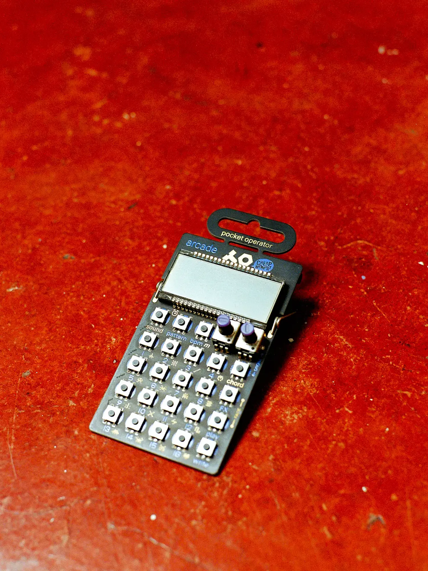 A small Teenage Engineering Pocket Operator PO-20 Arcade synthesizer resting flat on a textured, vibrant reddish-orange floor. The micro-device features an LCD screen, an array of grid-style button switches, and two small knobs.