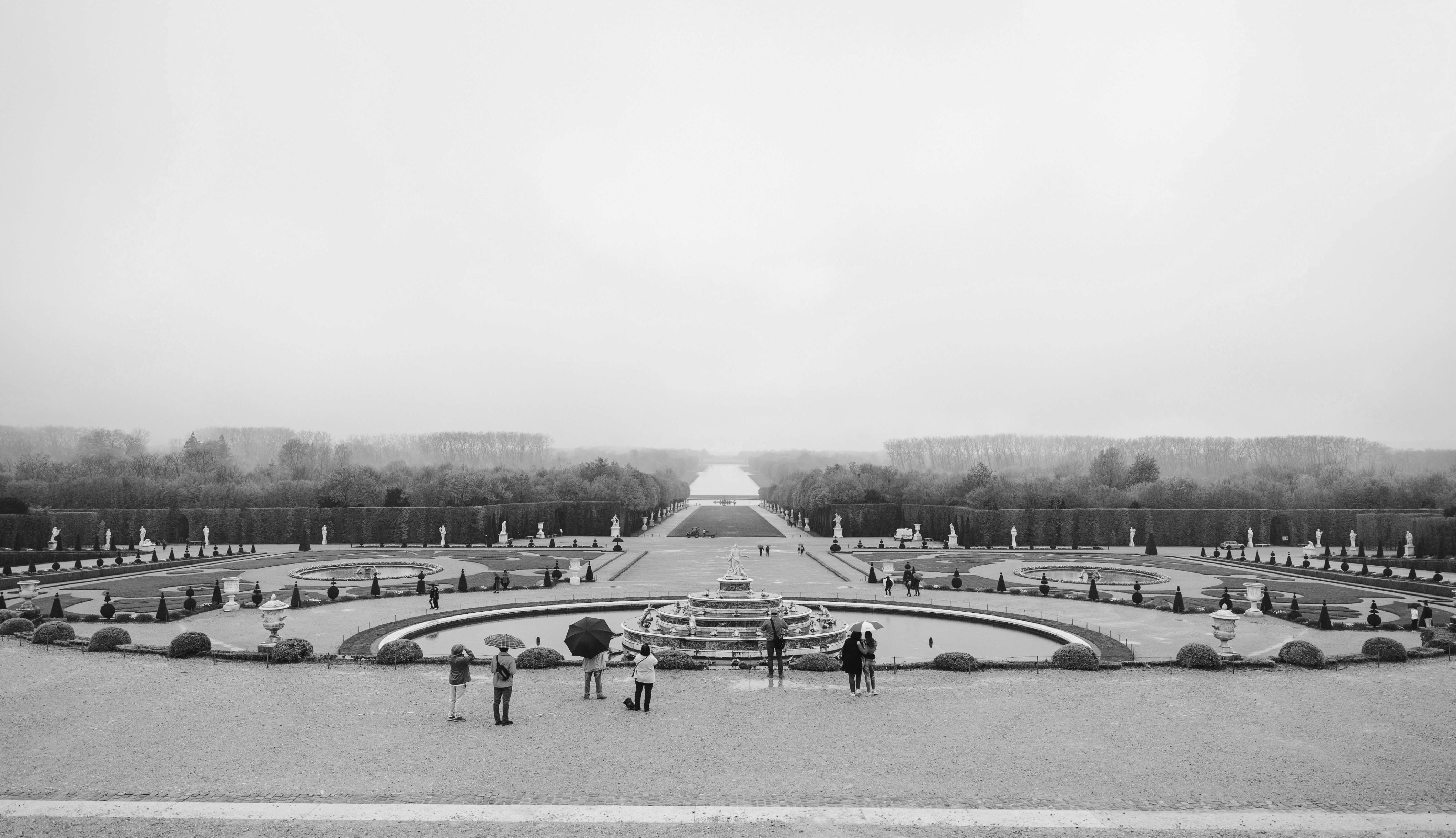 A black and white photo of the Versailles garden with a tiered fountain and a canal in the background.