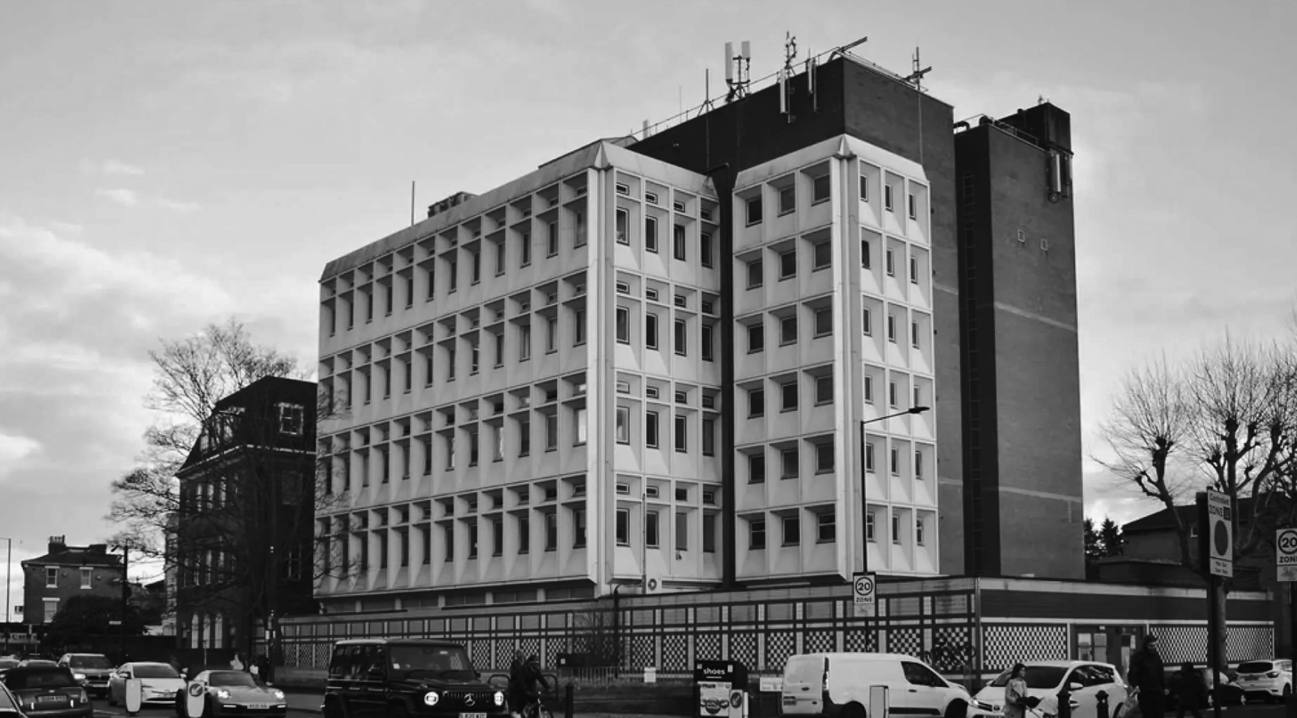 A black and white photo of a large, boxy building with many windows. The building is several stories tall and has a flat roof. There are trees and other buildings in the background.