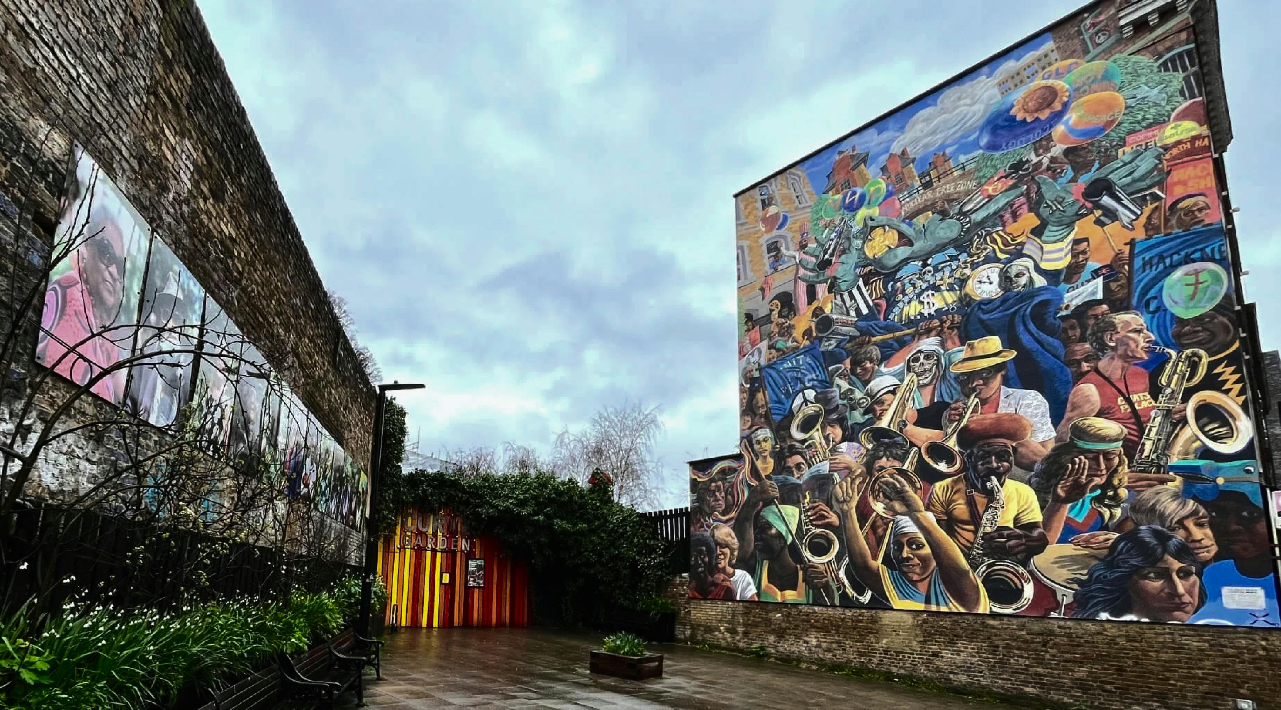 Brick building with a large mural depicting a jazz band. The mural is in the center of the image, and the building is on the right side. There is a brick wall on the left side of the image, and a tree in front of it.